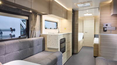 Interior of the Adria Altea Tyne with grey sofas, cosy cushions and wood accents, illuminated by natural light through windows, offering a view outside.  Perfect for relaxation. and designed for comfort.