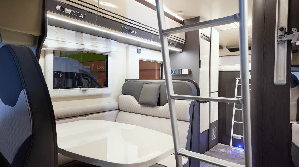 The Roller Team Zefiro 675 has grey carpet.  The upholstery is cream with dark brown highlights.  There is a cream table in front of the sofas.  The cabinets are cream with dark brown surrounds.  There are two ladders to gain access to beds.