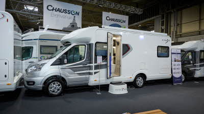 Auto-Trail F-Line F72's exterior is white with a silver cab.  There are grey and silver decals.  The entrance door is open revealing the lounge and there is a white step to gain easy access.  There is a roller banner to the rear.