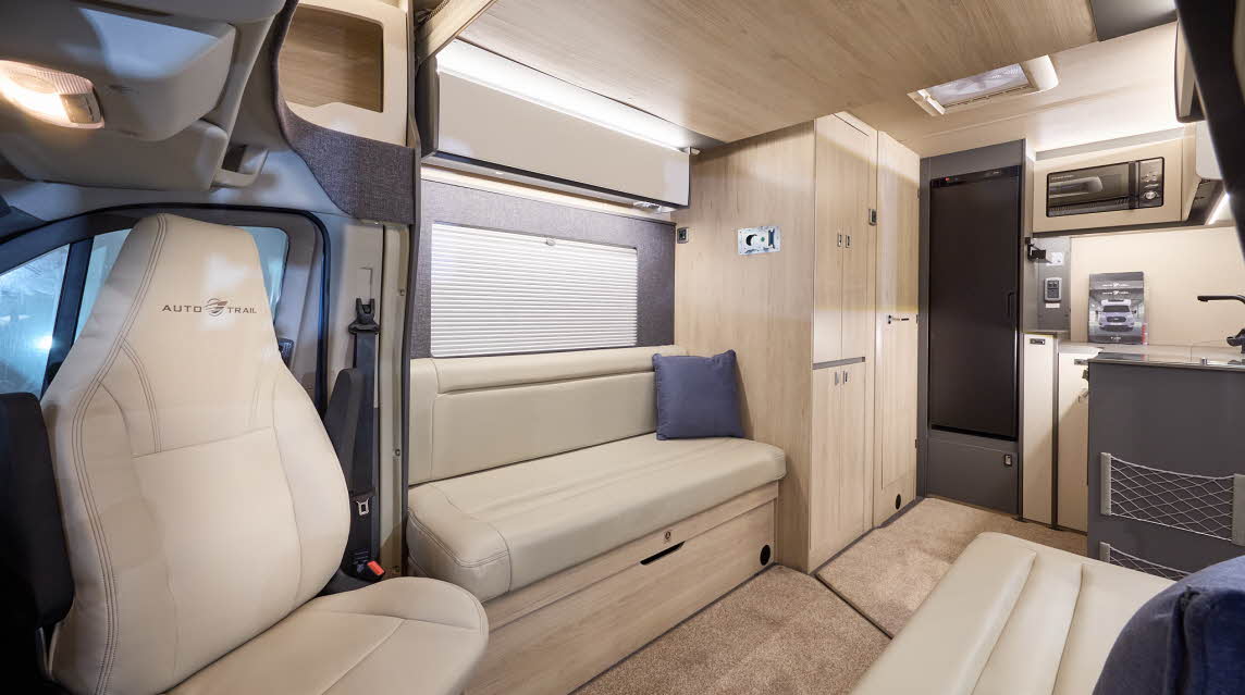 The Auto-Trail F-Line F62 has beige carpets, with pale wood cabinets with cream overhead lockers.  The upholstery is cream leather.  There is a rear kitchen with a black fridge freezer and microwave in an overhead cabinet.