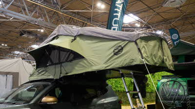 The Latitude Tents Pioneer is displayed on a green Dacia vehicle.  It has green canvas walls and a pale grey roof.  It has an extended platform with silver ladder to gain easy access.
