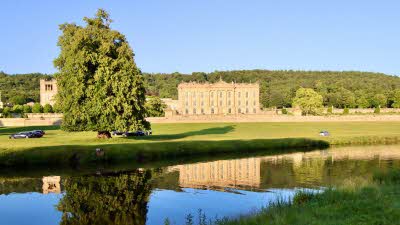 Chatsworth House sits in green fields surrounded by trees against a blue sky, seen across the lake 