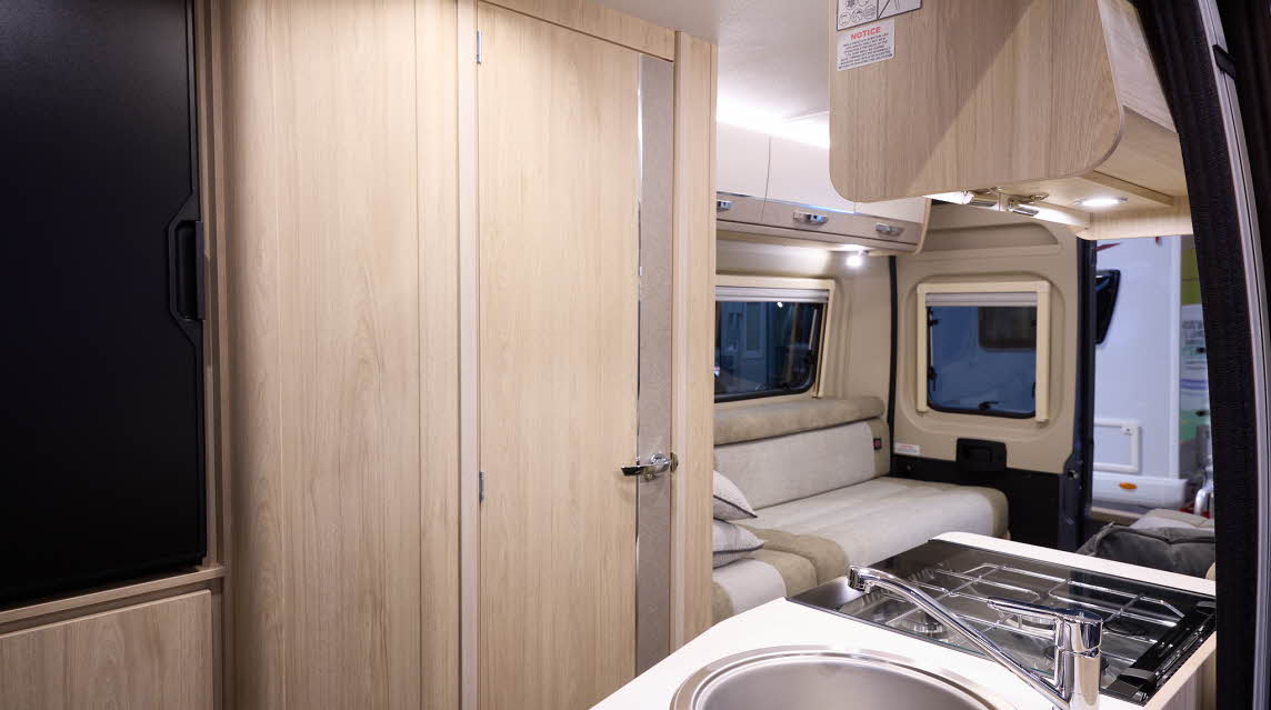 The Elddis Autoquest CV20 has cream and pale brown cabinets.  The upholstery is cream and pale brown with dark brown piping.  The round sink is in the front.  There is a rear lounge with two sofas. One of the rear doors is open.