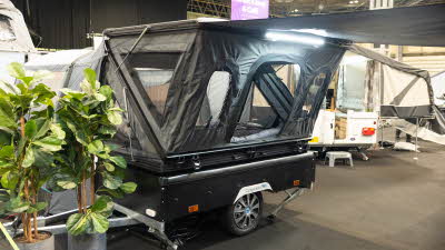 The Campmaster One Motion has a black base and its canvas is also black.  There are many windows.  There are two blue chairs in the extended canopy.