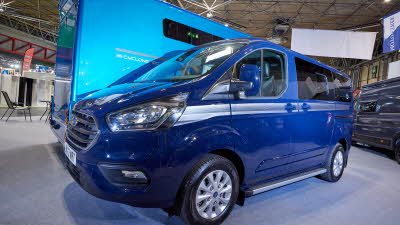 Three Peaks Ford Custom Campervan has a blue exterior and has a silver horizontal strip.  There is a sliver step into the vehicle.  The wheels are silver alloys.