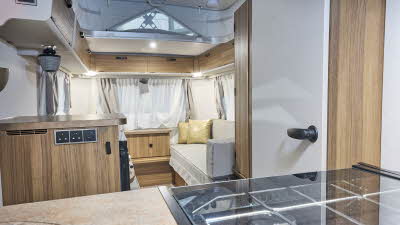 Interior of the Eriba Touring 420 Legend with cream sofas, cushions, wooden accents and a wooden floor.  To the front is the kitchen and its pop top roof is open.