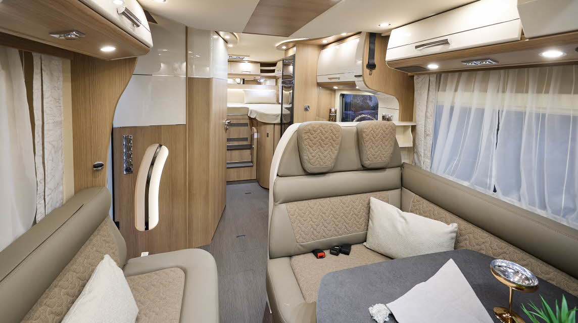 Carthago c-tourer T 148 LE H’s interior has beige leather upholstery with fabric panels.  The furniture is wooden with cream doors.  Nets curtains hang at the windows.  There is a fixed bed to the rear.  