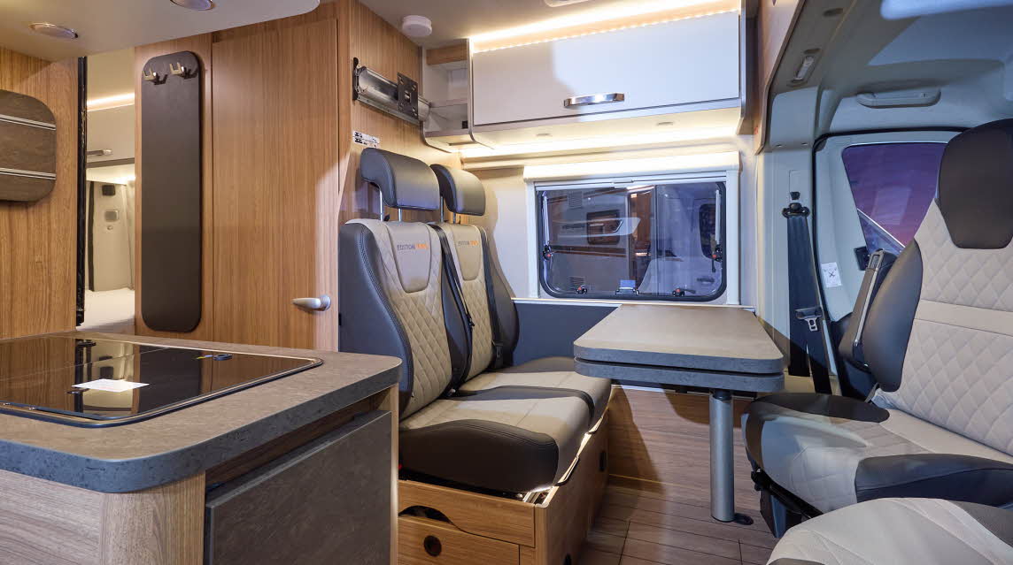 The Weinsberg CaraBus 600 MQ Edition FIRE has a wooden floor and its upholstery is coffee and cream leather.  The cabinets are white with silver handles.  The furniture is wooden.  There is a hob and sink under the cover to the front left.