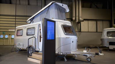 Eriba Touring 620 Legend’s exterior is silver.  It’s extended pop top roof is up.  There are two wooden steps to gain easy access and there’s a large interactive screen to the right.