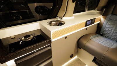 The Hillside Leisure MVX has black and blue upholstery, its cabinets are pale wood. There is a single gas hob ring and a round stainless steel sink.