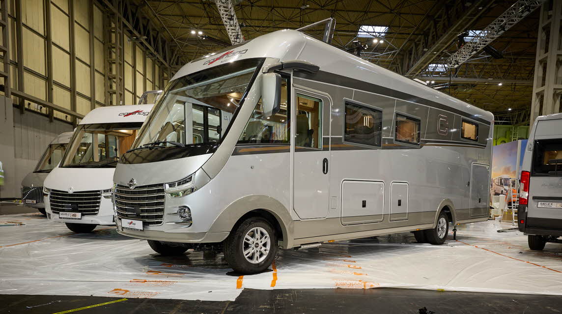 Carthago chic s-plus I 64 XL QB’s exterior is silver.  It’s entrance door is shut.  There are three external lockers whose doors are closed.