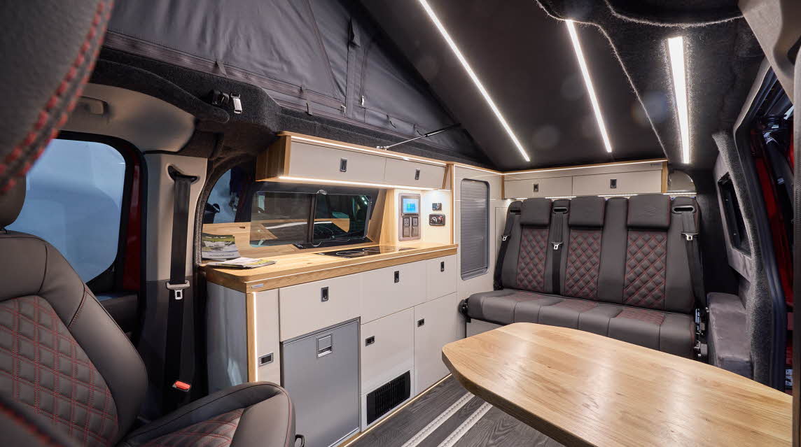 The Landseer Artisan Red Ford Custom 2.0 has a large wooden table and its upholstery is black with red stitching. The cabinets are cream and pale wood. The rising roof is fully open.