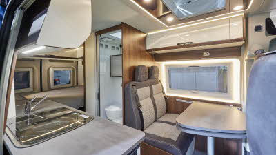 Globecar Summit Prime 600 has two tone grey and beige upholstery, cream and wooden overhead lockers.  The kitchen work surface is in grey with the central washroom opposite.  There is a fixed bed at the rear.