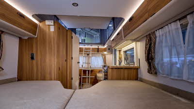 Eriba Touring 642 Legend's interior has wooden furniture with grey upholstery.  The fixed bed is at the front of the photograph.  The pop top roof is open with a ladder for easy access.