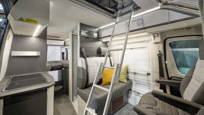 Adria Twin Sport's interior has brown and beige upholstery with two yellow cushions, there is a silver ladder to the roof bed and the driver’s and front passenger's seat are swivelled.  The fixed bed is to the rear with the hob on the left hand side