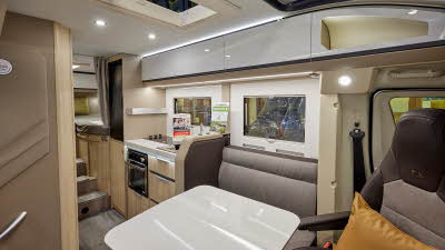Adria Compact Supreme DL has grey upholstery, with shiny white overhead cabinets and table.  It has light wooden panelling, there are mid-brown carpets, there are three steps leading to the fixed rear bed with a privacy curtain.