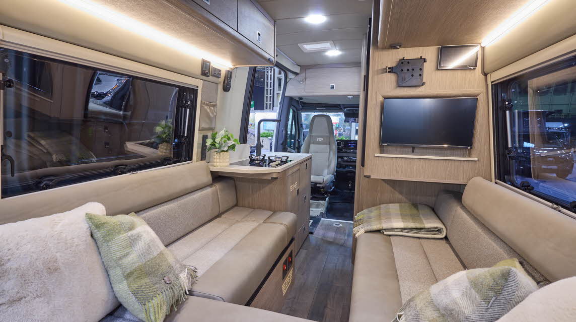 The RP Overland Rebel has wooden flooring. The upholstery is cream with a matching pattern. The cabinets are wooden. There is a television on the right hand wall. The passenger seat is facing into the vehicle. There is a u shaped lounge.