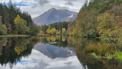 A image of Glencoe from the waters edge looking out over the still loch water reflecting the high green trees on either side and the high mountain rising in the distance under a sky covered in white clouds with some hints of the blue beyond