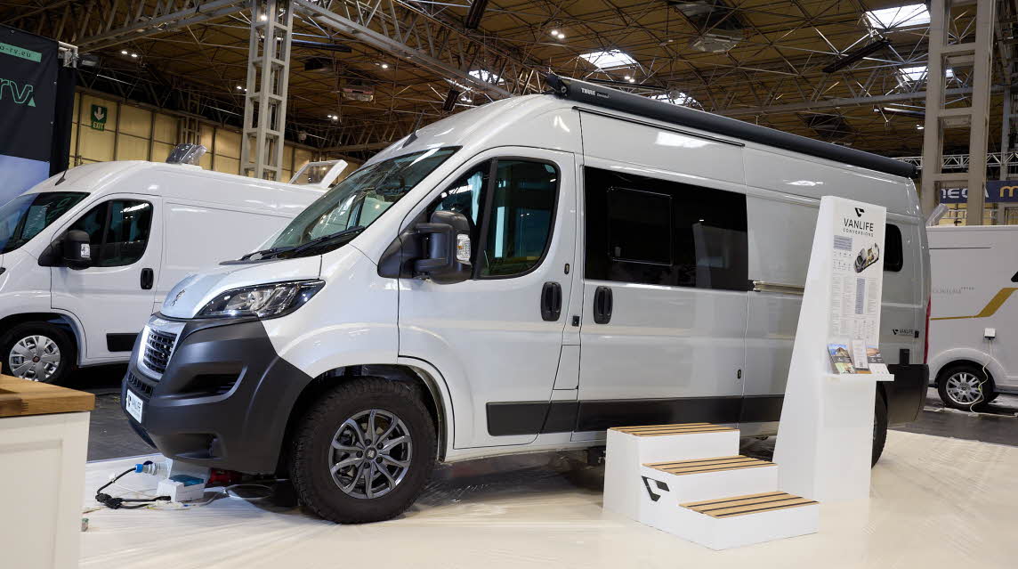 Vanlife Conversions Kunu is silver with black trim.  There are three steps outside the closing sliding door  There is an infomation stand next to the steps.