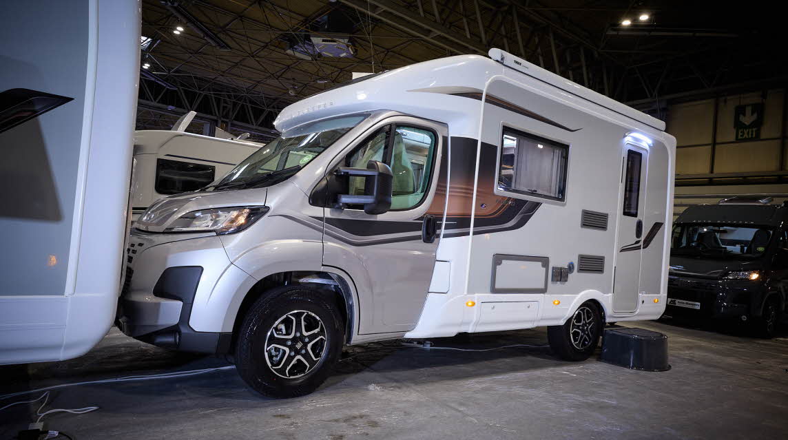 The Auto-Sleeper Nuevo has a gold cab with a white exterior with a grey inset. It has black/grey decals. There is a black step by the habitation door.