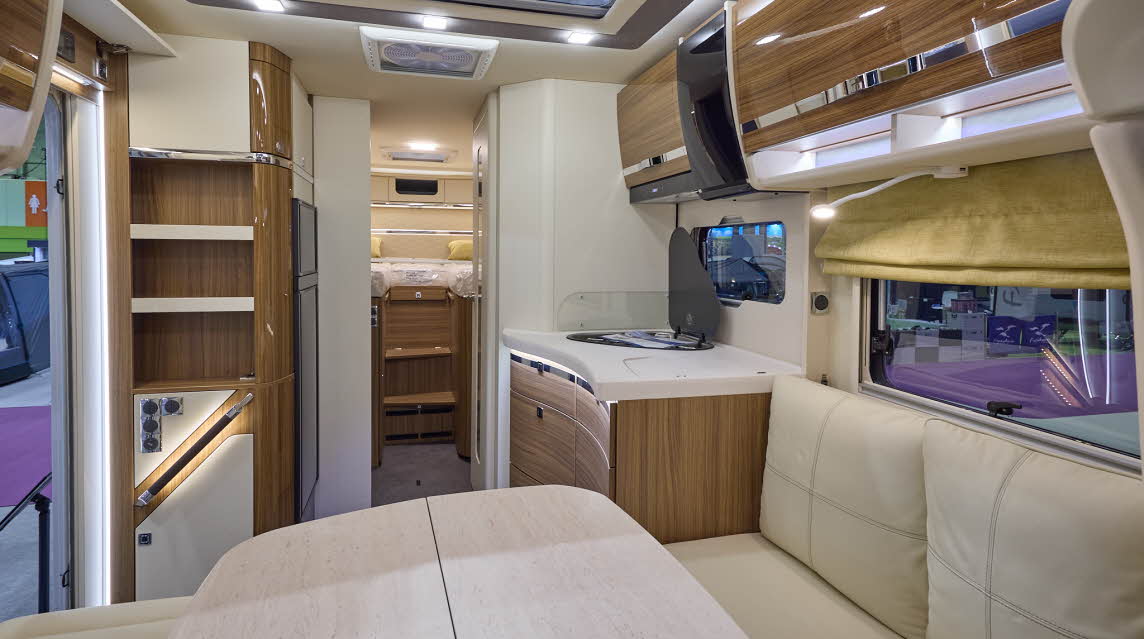 The Eura Mobil Contura CT 766 EB has grey carpets and shiny wooden cabinets.  The upholstery is cream leather.  There is a large cream table in the front.  There are two steps to the rear fixed bed.