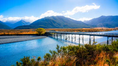 Bridge crossing over a calm river with huge mountains in the distance topped with fluffy clouds under a sunny blue sky