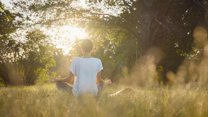 a woman in comfortable clothing sits cross-legged facing the sunrise surrounded by trees