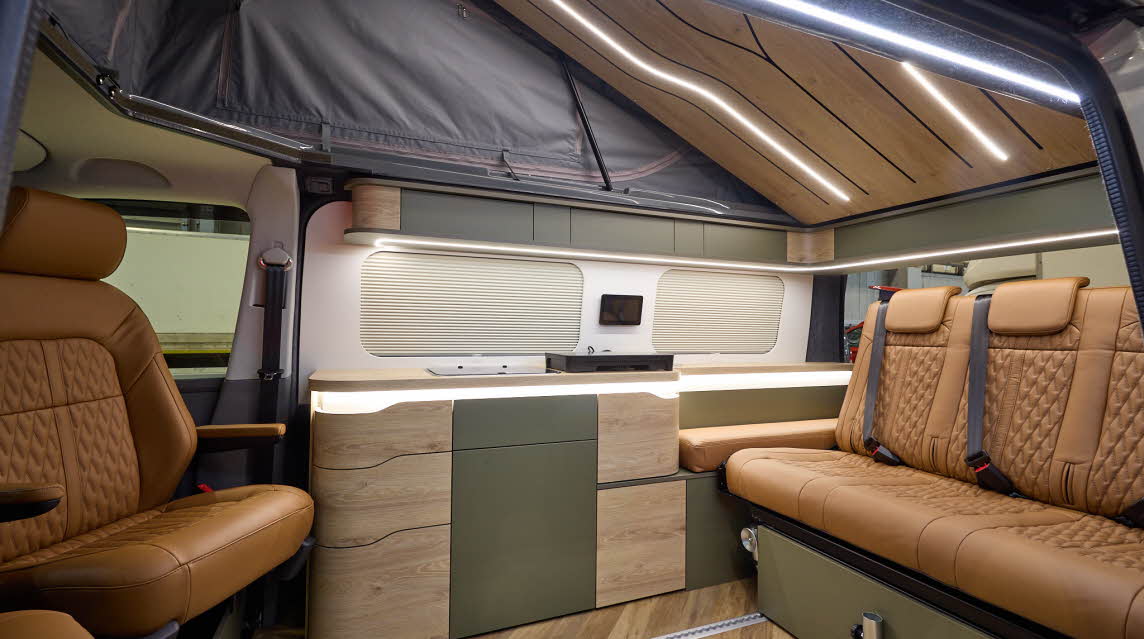 The Sherwood Campers Ltd Loxley Edition Conversion has wooden parquet-style flooring and its upholstery is brown leather. Its furniture is wooden which is curved. There are dark green inserts. The rising roof is open, there's strip lighting.