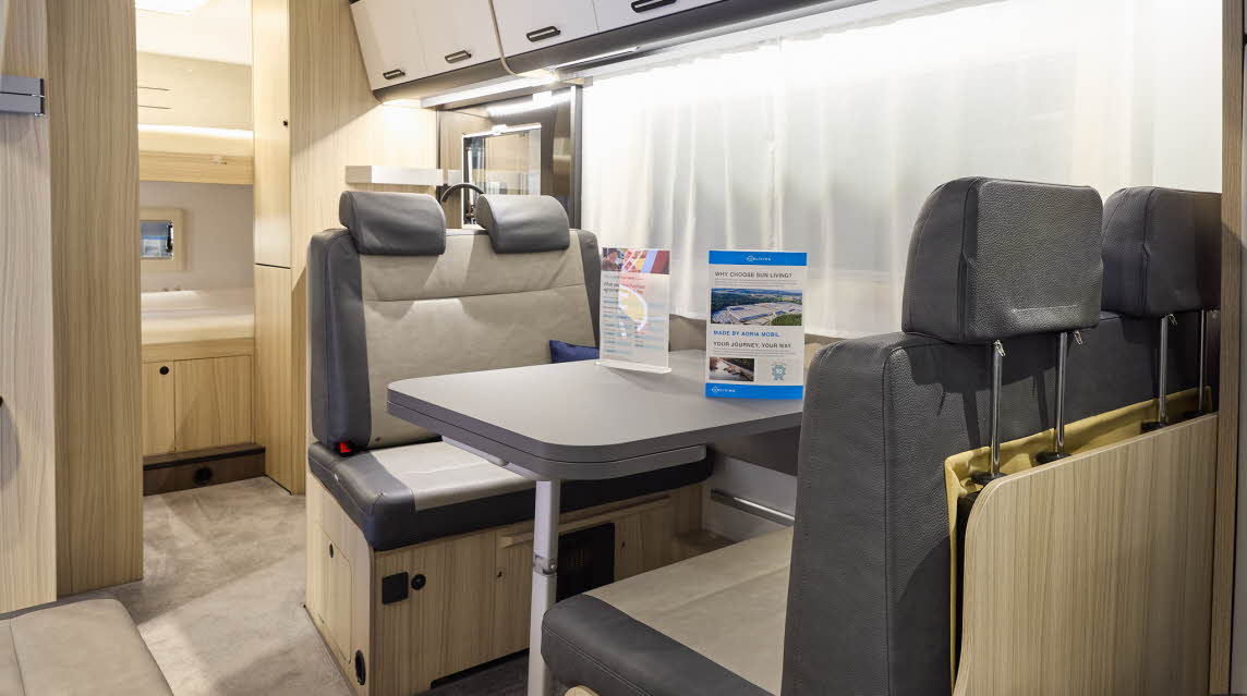The Sun Living A Series 70DK has beige carpet and its upholstery is dark brown with cream inserts. The cabinets are white and dark wood. Towards the rear there are fixed bunks.