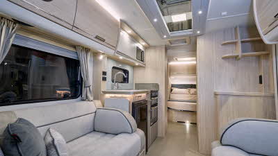 Elddis Crusader Aurora’s interior has grey upholstery with dark grey accents.  It has pale wooden furniture.  The kitchen is in the middle and the bedroom door is open revealing a fixed island bed.