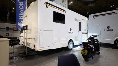 The Rimor Sarus 66 Plus is white with black and bronze decals.  There are three white steps with a blue cover in front of the habitation door.  There is a motorbike parked outside the vehicle.
