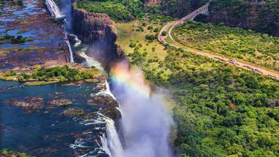 rainbow over the Victoria Falls, with water on the left cascading over the edge of the crevasse and lush green forest the other side