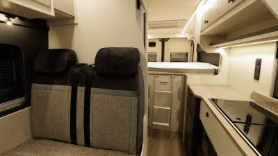 The Ace Motorhomes 1200 GL has brown and fawn upholstery.  It has cream cabinets with square handles.  There is a rear fixed bed which has a silver ladder to gain access.