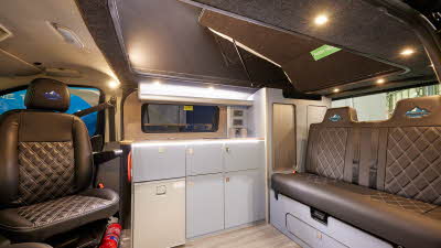 Three Peaks Ford Custom Campervan brown embossed upholstery.  The cabinets are a pale grey and it has a cream fridge.  The floor is wooden laminate.  Under the swivelled driver’s seat is a fire extinguisher.  The rising roof is closed.