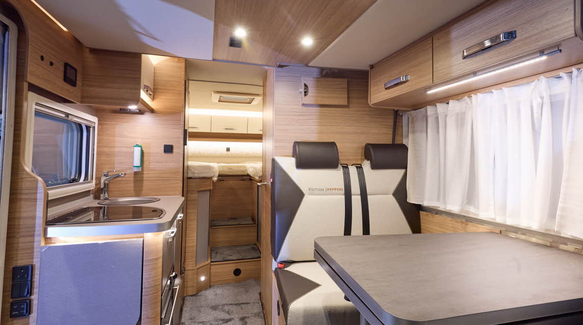 The Weinsberg CaraCompact Suite MB 640MEG Edition Pepper has a grey carpet, its upholstery is coffee and cream.  The furniture is wooden with silver handles.  There are two steps to the fixed rear bed.