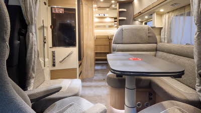The Malibu Mercedes-Benz T 450 RB-LE lightweight 3.5t has grey flooring with cream and wooden cabinets.  The upholstery is beige and grey.  There is a grey table in front of the sofas.  There are two steps to the fixed rear bed.
