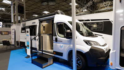 The Sun Living V Series 65 GX has a white exterior with black trim.  Its exterior door is open and there are two black steps to gain easy access.  There is a display stand next to the steps