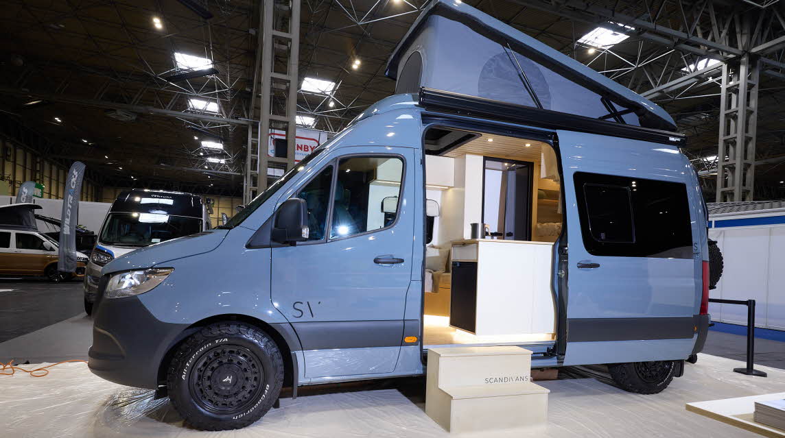 The Scandivans Sprinter 907 MWB L2H2 has a pale blue exteriors.  There are two steps leading to the open sliding door revealing the interior.  Its rising roof is fully extended.