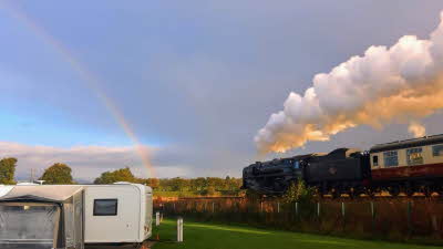 Steam train with white smoke blowing from the smokestack alongside a caravan pitched on green grass next to the track, with sky and a faint rainbow in the distance