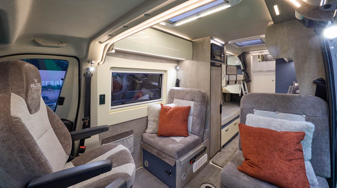 WildAx Motorhomes Solaris XL has brown and cream upholstery with two cushions on each passenger seat.  It has beige carpet.  To the rear there are fixed bunks with a silver ladder.