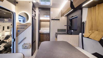 The Rimor Sarus 66 Plus has a cream floor.  The upholstery is cream, and it has gold curtains.  The cabinets are all wooden and the interior door is a dark brown.  There is a fixed rear bed.  The vehicle has a large table in front of the sofas.