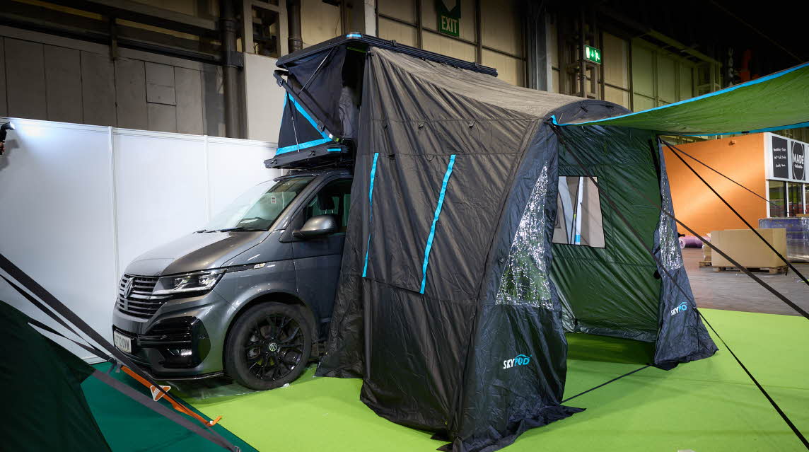 The SkyPod Lair is displayed on a Volkswagen campervan.  It has grey canvas walls and a black hard roof.  It has turquoise flashes around it.  It has a matching annex and awning.