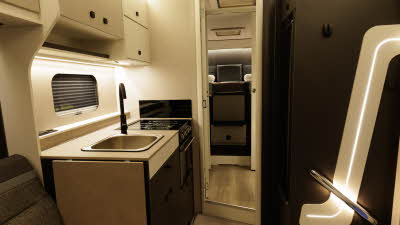 The Ace Motorhomes 1500 SL has pale wood and white cabinets.  The entrance has a strip light and grab rail.  The kitchen has a square sink and a full size oven.  There is a work top extension flap beside the sofa.  At the rear there's two single beds