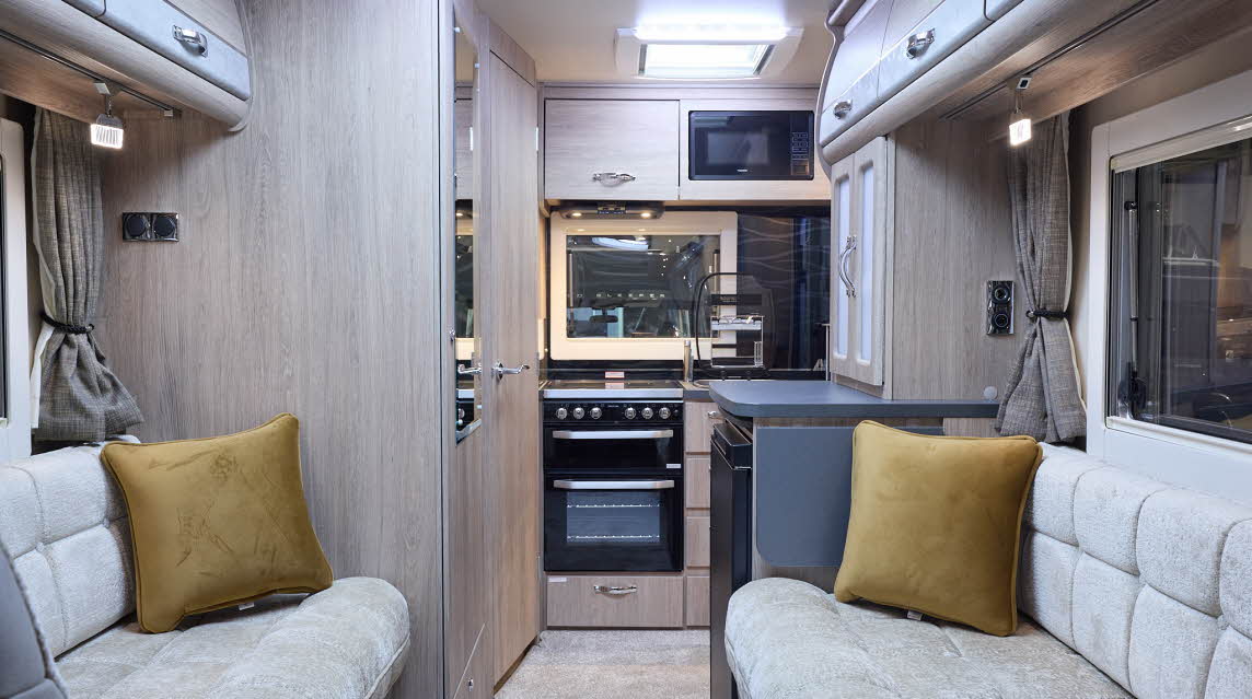 The Auto-Sleeper Nuevo has cream carpet, with pale wood cabinets with pale grey highlights. The upholstery is cream. There are two gold cushions. The kitchen is at the rear with an oven and the sink cover is open.