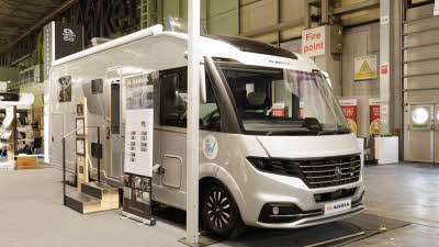 The Adria Supersonic 780 DL is silver.  There are two steps beside its habitation door.  There is a roller banner one side of the steps with an interaction stand on the other side.  Its skylights are open.