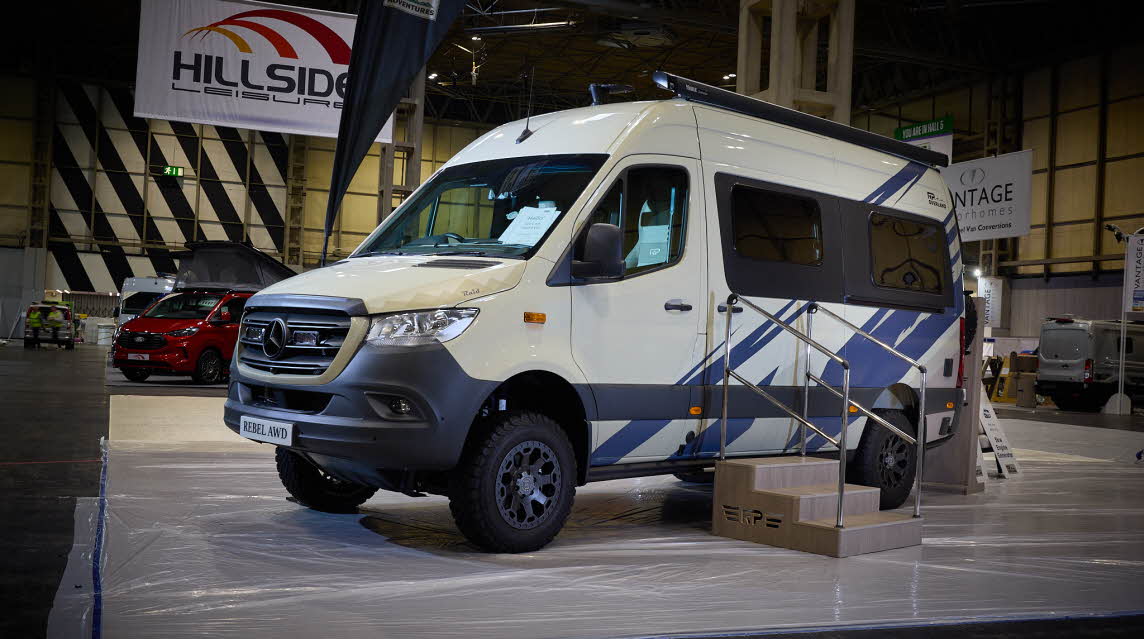 The RP Overland Rebel Raid AWD has a cream exterior with striking blue decals. There are three steps leading to the closed sliding door.