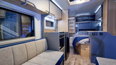 Bailey Discovery D4-4 interior, it is beige with light wood and blue accents.  The kitchen is in the centre with a fixed bed just off it.  The washroom is at the rear.  It has a skylight which is open.