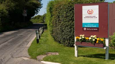 A sign beside the road for Blackpool South UK Club Campsite with flower pots full of lovely yellow flowers