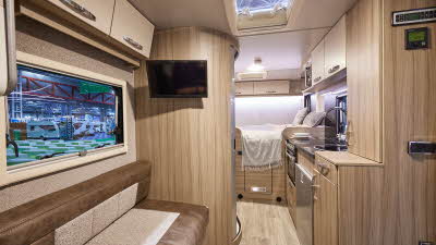 Moto-Trek Ltd Xplora FDB has two tone beige and brown upholstery. The furniture is wooden with two tone cream and brown overhead lockers.  There is a TV mounted on the wall of the washroom.  Towards the rear of the vehicle there is a fixed double bed