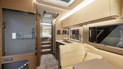 Adria Supersonic 780 SL interior which has cream upholstery, with cream overhead cabinets and wooden curved panelling.  The carpet is mid brown, there are three steps leading to the fixed rear bed.  There is a large roof light above the kitchen.